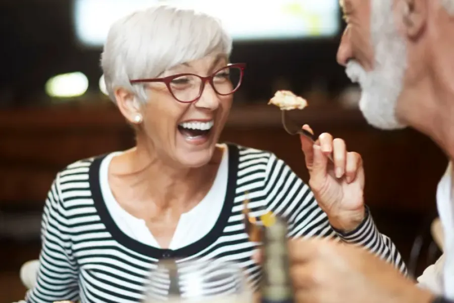 Acclaim at the Hill senior apartment homes with Older couple laughing and sharing food at a restaurant, enjoying drinks and a meal together.