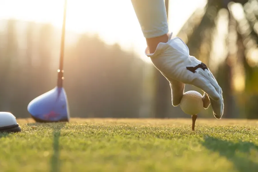 Acclaim at the Hill senior apartment homes with Gloved hand placing a golf ball on a tee at sunrise, with a golf club and grass visible.