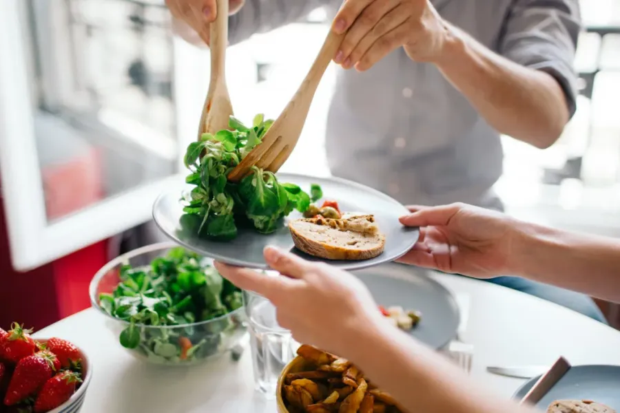 Acclaim at the Hill senior apartment homes with Hands serving salad onto a plate beside strawberries, bread, and other dishes on a white table.
