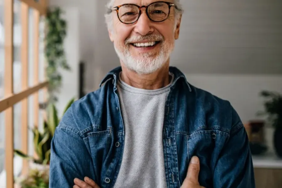 Acclaim at the Hill senior apartment homes with Smiling older man with glasses and gray hair, wearing a denim shirt, standing by a window indoors.