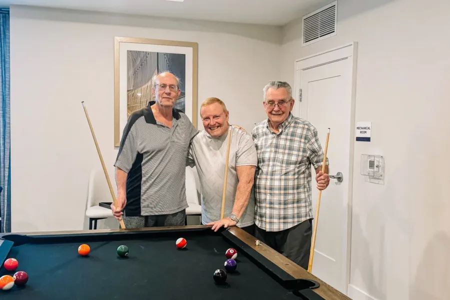 Acclaim at the Hill senior apartment homes with Three older men smiling and posing with pool cues around a pool table in a bright, modern room.