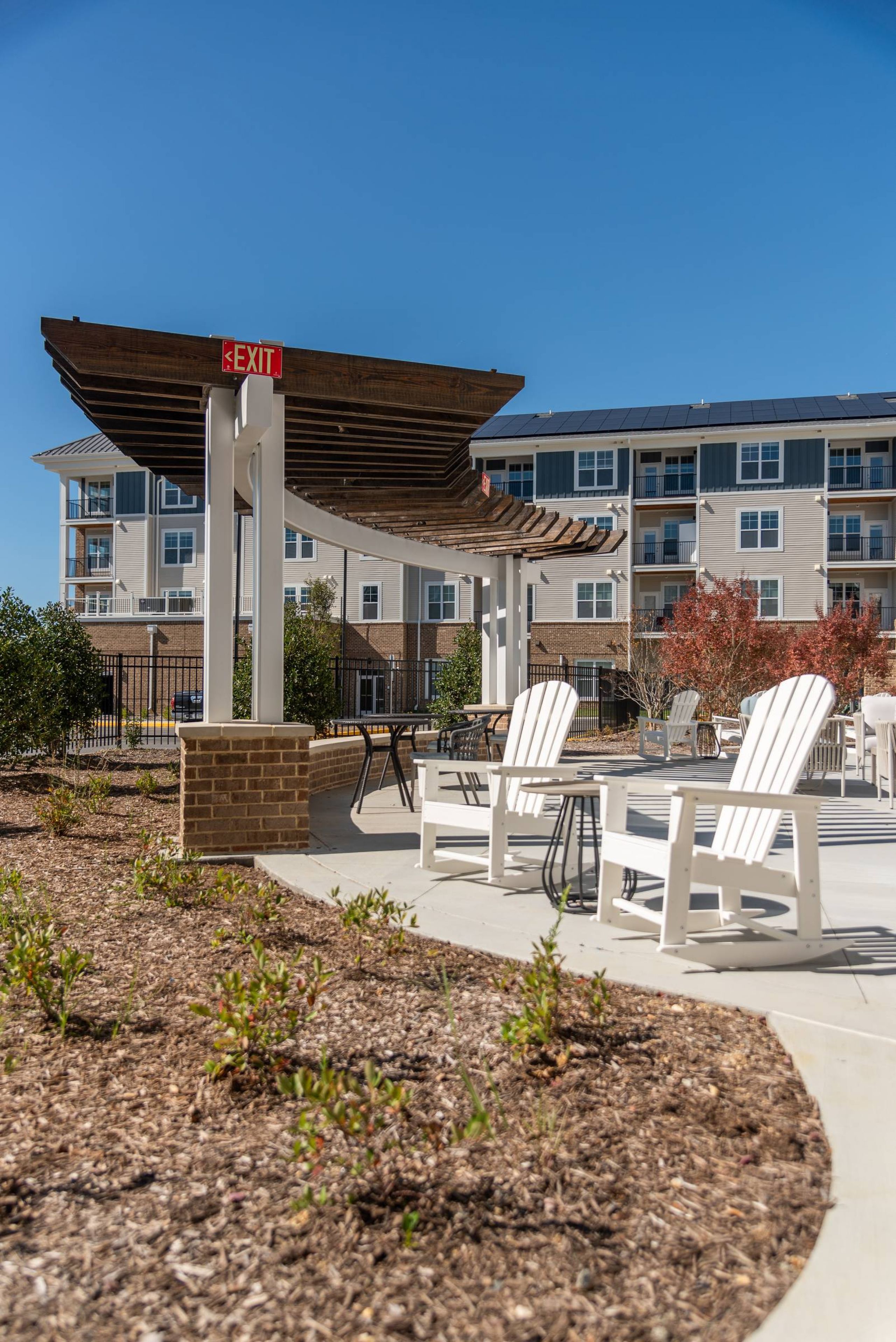 Acclaim at the Hill senior apartment homes with White chairs sit on a patio under a pergola near apartment buildings on a sunny day.