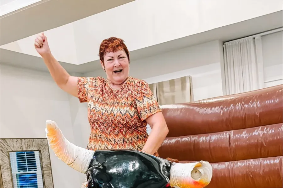 Acclaim at the Hill senior apartment homes with Smiling woman rides a mechanical bull indoors, raising one arm in excitement.