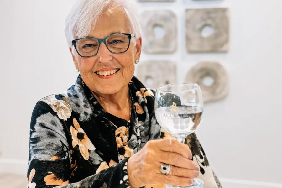 Acclaim at the Hill senior apartment homes with Smiling older woman with gray hair holds up a glass of water, wearing glasses and a floral top.