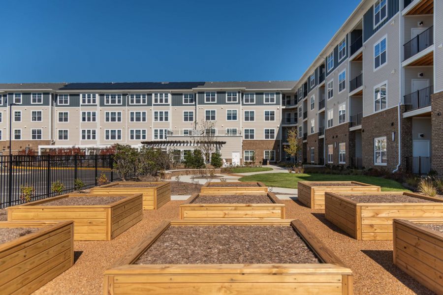Acclaim at the Hill senior apartment homes with Raised garden beds in front of a modern apartment building with balconies on a clear, sunny day.