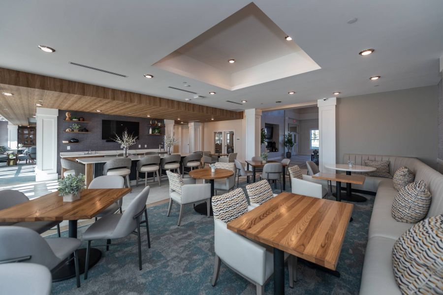 Acclaim at the Hill senior apartment homes with Modern lounge with wooden tables, gray chairs, cushioned benches, and a bar area in the background.